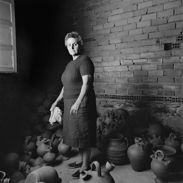 Agusti mirando de frente, triste, rodeada de piezas de barro y con un paño para sacudir los botijos en su mano